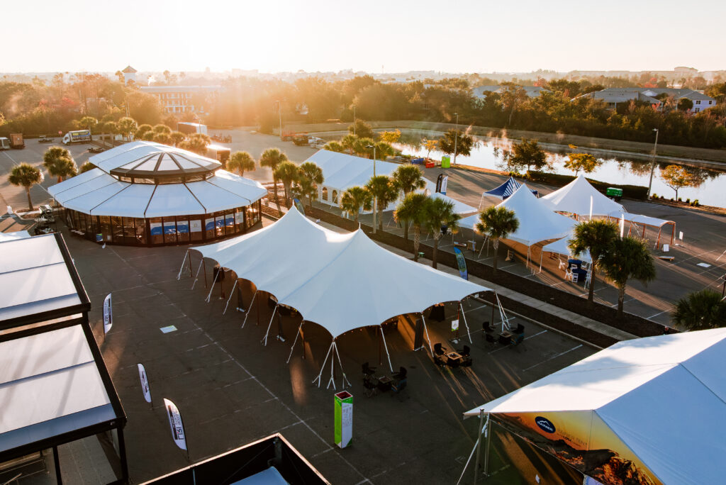 Spacious outdoor venue featuring white tents and a circular building, bathed in warm sunlight, near a waterway.