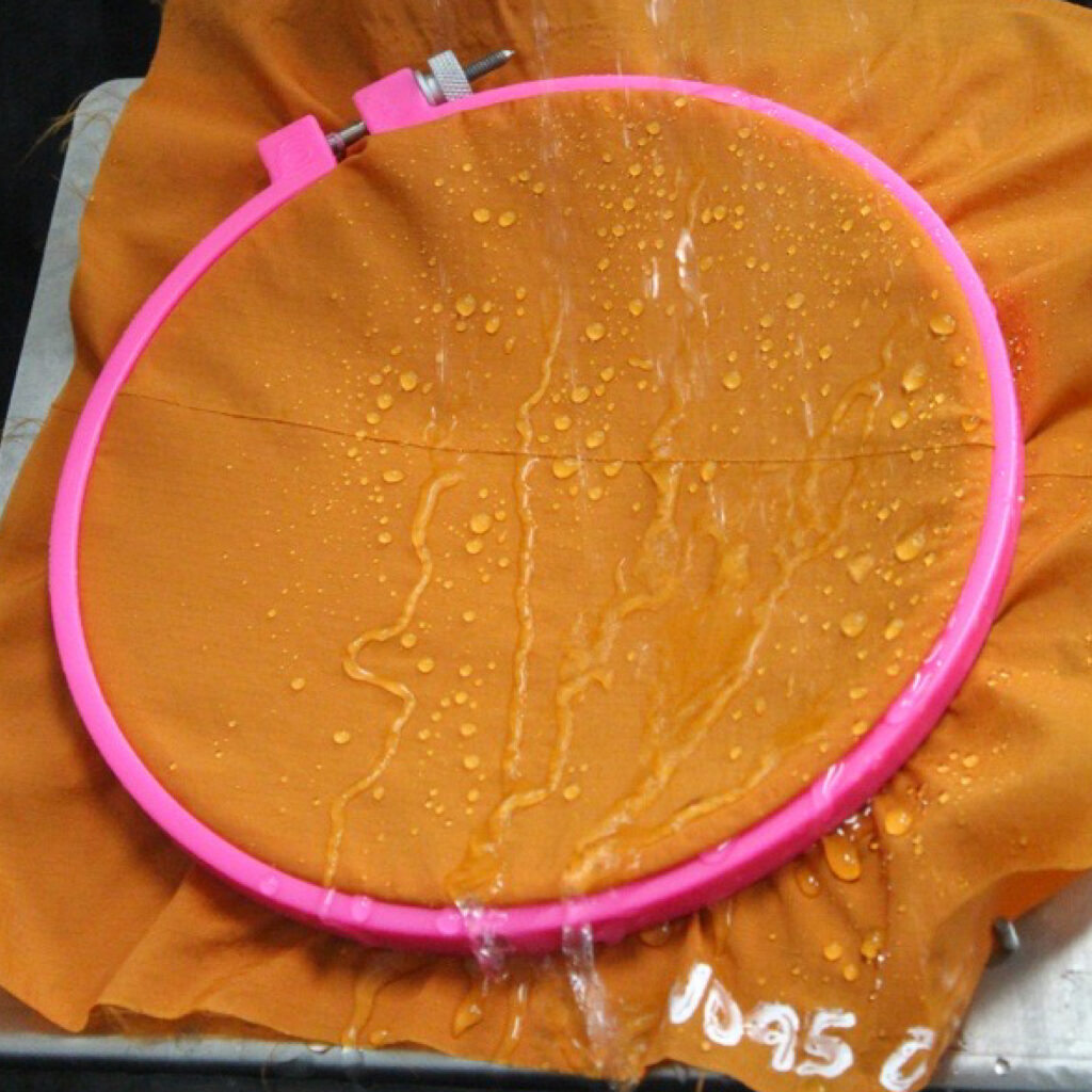 Water droplets bead on an orange fabric secured in a pink embroidery hoop.