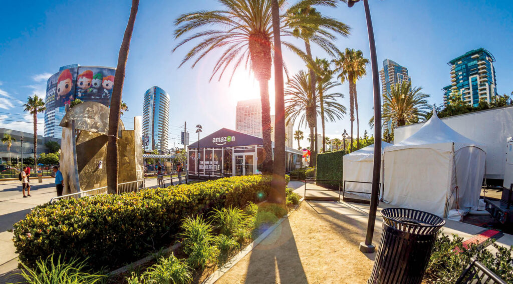 A vibrant outdoor scene featuring palm trees, an Amazon booth, white tents, and modern buildings under a bright blue sky.