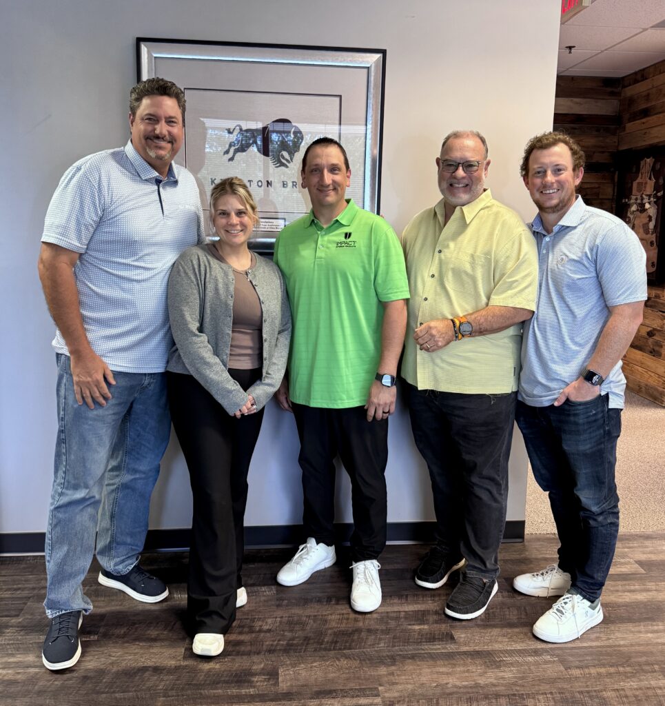 Five smiling individuals stand together in an office setting, wearing casual attire in various colors. A framed photo is displayed behind them.