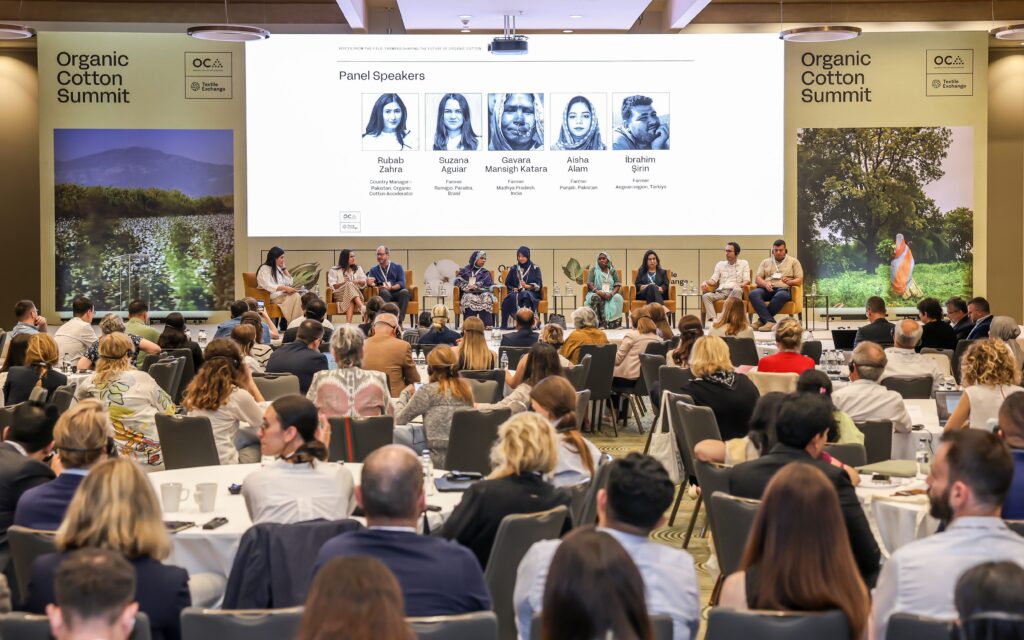 A panel discussion in a conference room, featuring diverse speakers at a long table, with an audience of attendees in the foreground.