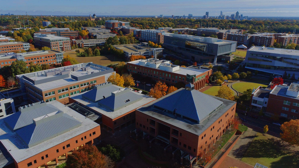 Aerial view of a university campus with modern buildings, colorful autumn trees, and a skyline in the distance under a blue sky.