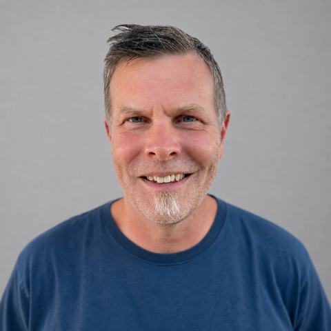 Smiling middle-aged man with short gray hair and a beard, wearing a blue t-shirt, against a light gray background.
