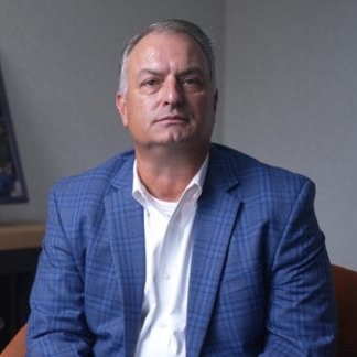 Middle-aged man with short gray hair, wearing a blue plaid blazer and white shirt, seated in an office setting with a neutral background.