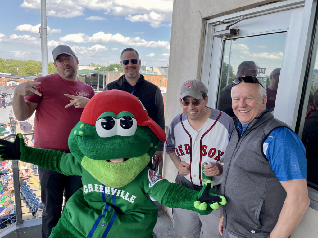 Five men pose together with a green frog mascot wearing a red cap and Greenville jersey, in a lively outdoor setting with blue skies.